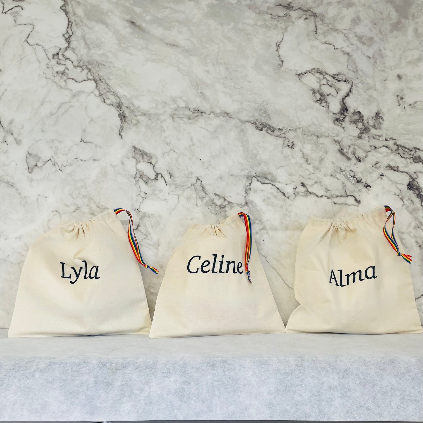 black lettering embroidered onto cotton drawstring bags with rainbow stripe ribbon handles.