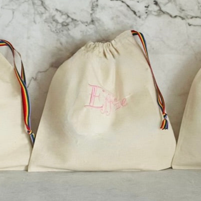 unbleached cotton drawstring bag embroidered in baby pink with the name Effie. A rainbow stripe ribbon drawstring is closing the bag.