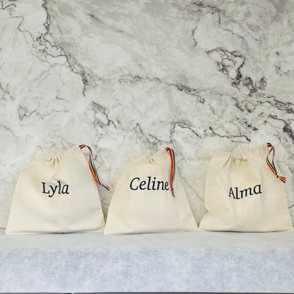 black lettering embroidered onto cotton drawstring bags with rainbow stripe ribbon handles. 