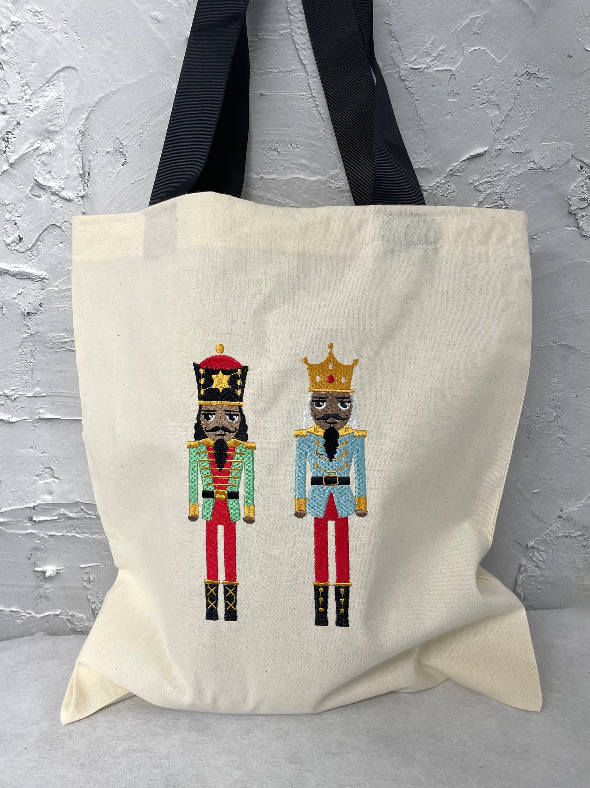 two large size nutcracker dolls wearing red trousers and green or blue jacket embroidered onto a natural canvas tote bag with black handle straps.
