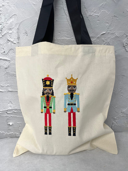 two large size nutcracker dolls wearing red trousers and green or blue jacket embroidered onto a natural canvas tote bag with black handle straps.